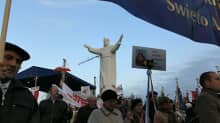 Ihmisiä patsaan paljastamistilaisuudessa, keskellä kuvaa näkyy kädet avoimeen kohotukseen nostanut Jeesus-patsas.