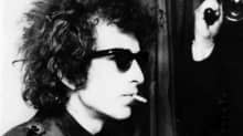 Bob Dylan tupakka suussa ja aurinkolasit silmillään. Taustalla mies kuvaa elokuvakameralla.