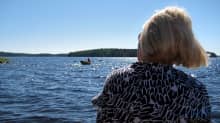Nainen (selin) istuu järven rannalla, katselee järvellä olevaa soutuvenettä.