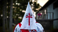 Ku Klux Klanin valkoiseen huppuun ja kaapuun pukeutunut mies tummassa illassa. Asussa on useita ristitunnuksia. 