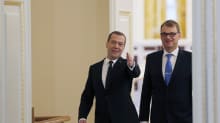 Venäjän pääministeri Dmitri Medvedev ja Suomen pääministeri Juha Sipilä tulevat suurista ovista. Miehillä on yllään mustat puvut, Sipilällä on sininen kravatti ja Medvedevillä tumma. Molemmat hymyilevät. Medvedev viittoo huoneen sisälle päin.