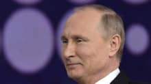 Putin hymyilee vienosti. Hänellä on tumma puku, valkoinen kauluspaita ja punainen kravatti. Taustakangas on tumma, siihen on kuvattu vaaleanvioletteja palloja.
