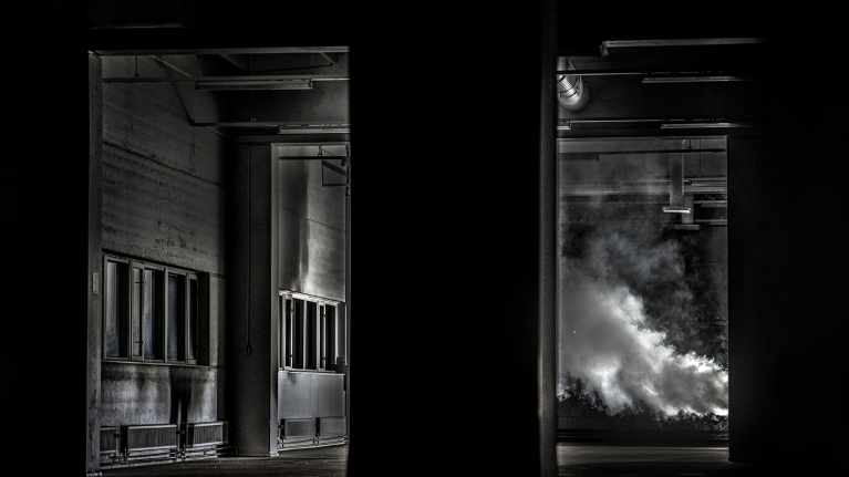 Tummassa tehdassalissa nousee savua.