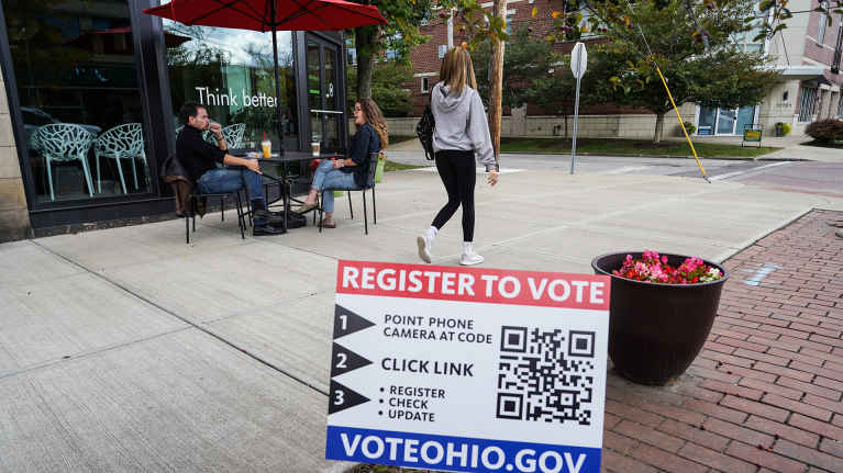 Register to vote -kyltti kuvan etualalla, taustalla katukahvila.