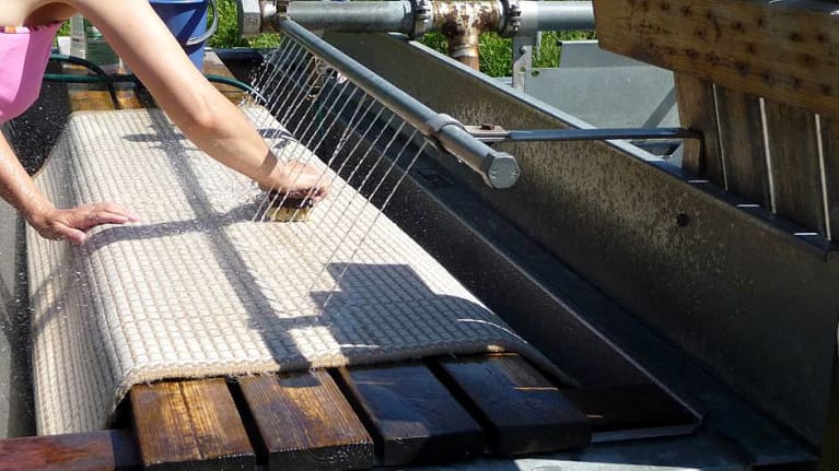 Nainen pesee juurharjalla mattoja matonpesupaikalla ulkona.