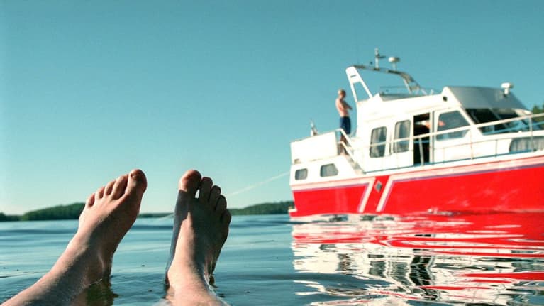 Miehen jalat veden pinnalla, taustalla punainen vene rantautuneena.