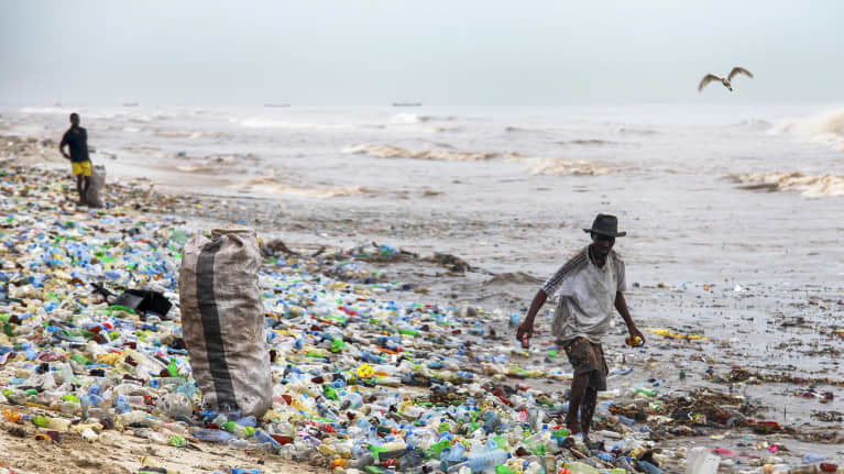 Kaksi miestä kerää purkkeja säkkeihin rannalla, joka on täynnä kirjavaa muovijätettä. 