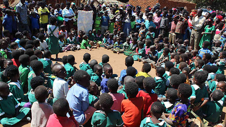 Lapsia istuu ja seisoo piirissä kuuntelemassa opettajaa. 