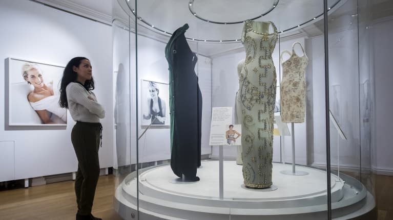 Mekkoja mallinukketorsojen päällä korokkeella, jota ympäröi läpinäkyvä seinä. Nainen seisoo vieressä. Takaseinällä on valokuvia prinsessa Dianasta.