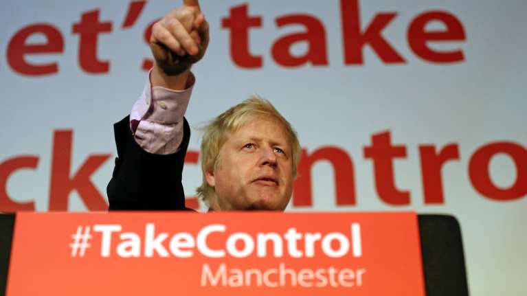 Boris Johson osoittaa puhujakorokkeelta sormella yleisöä. Puhujapöntössä on teksti, joka kehottaa ottamaan vallan itselle. 