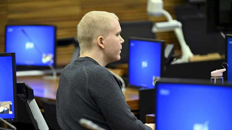 Aleksanteri Kivimäki istuu oikeussalissa ympärillään sinisiä monitoreja.