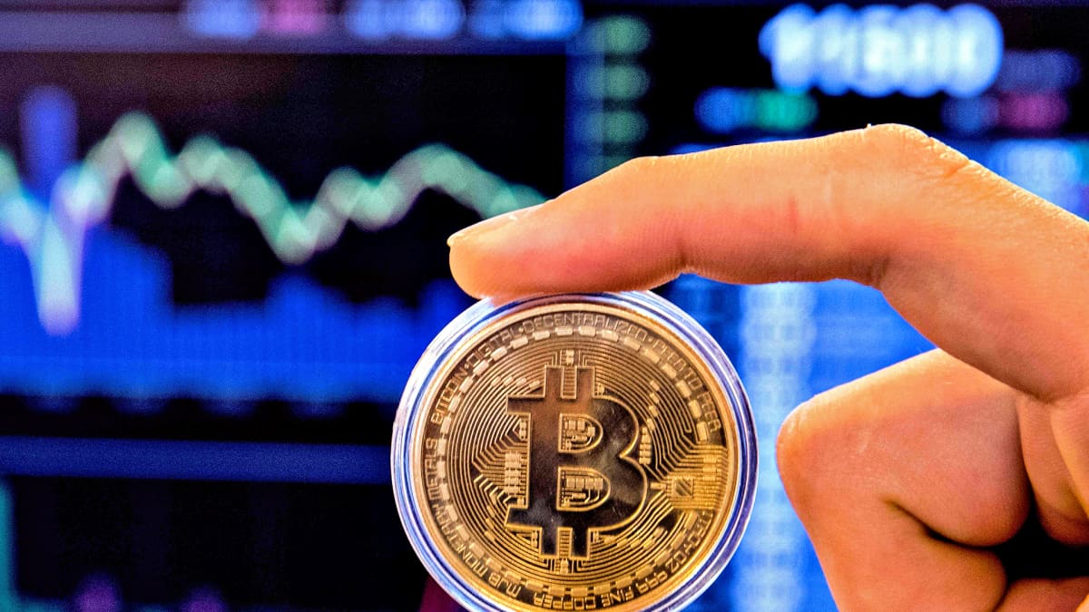 Käsi pitelee Bitcoin-kolikkoa. Takana näkyy alas kaartuva pörssikäyrä.