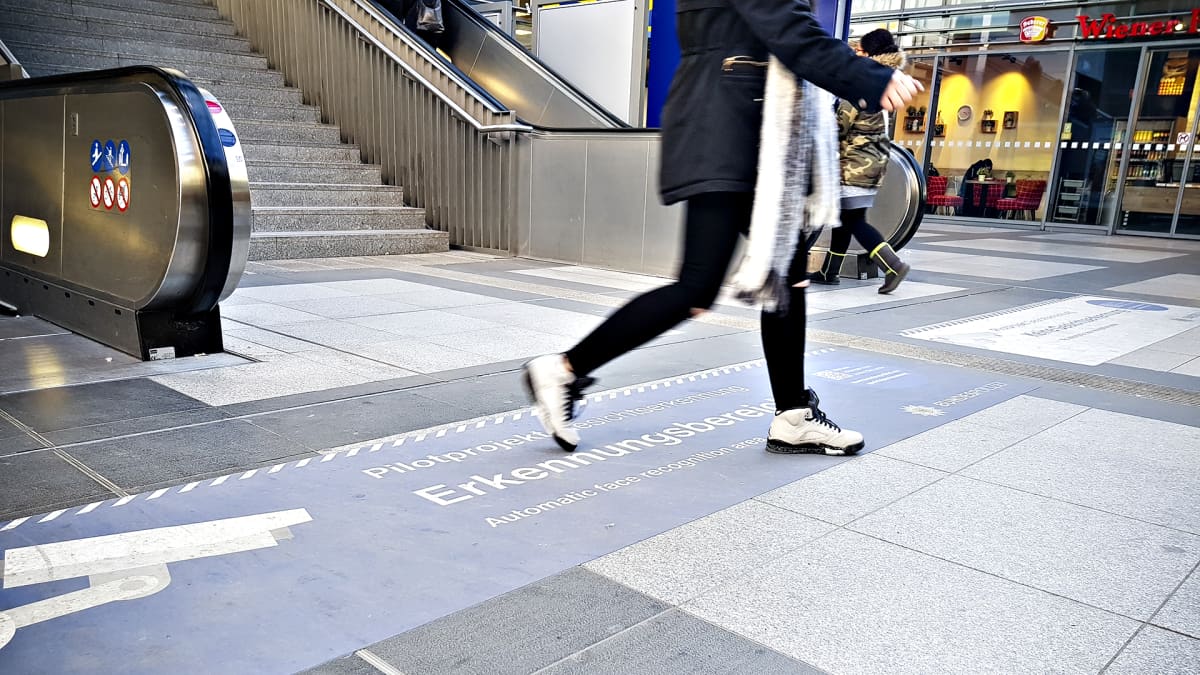 Suuret tarrat Berliinin Südkreuz-aseman lattiassa kertovat, mistä kasvojen tunnistusalue alkaa.  Nainen kävelemässä tarran yli.