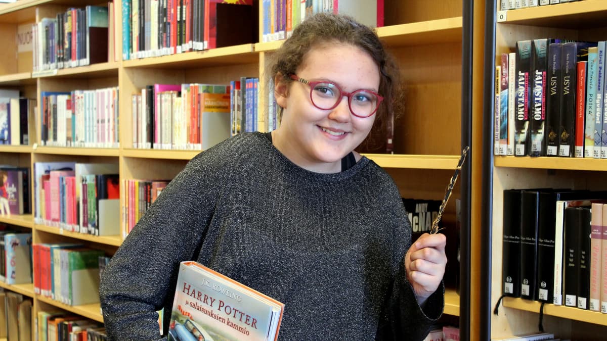 Aida Kankaanpää kirjastossa taikasauva kädessään ja Harry Potter -kirja kainalossaan.
