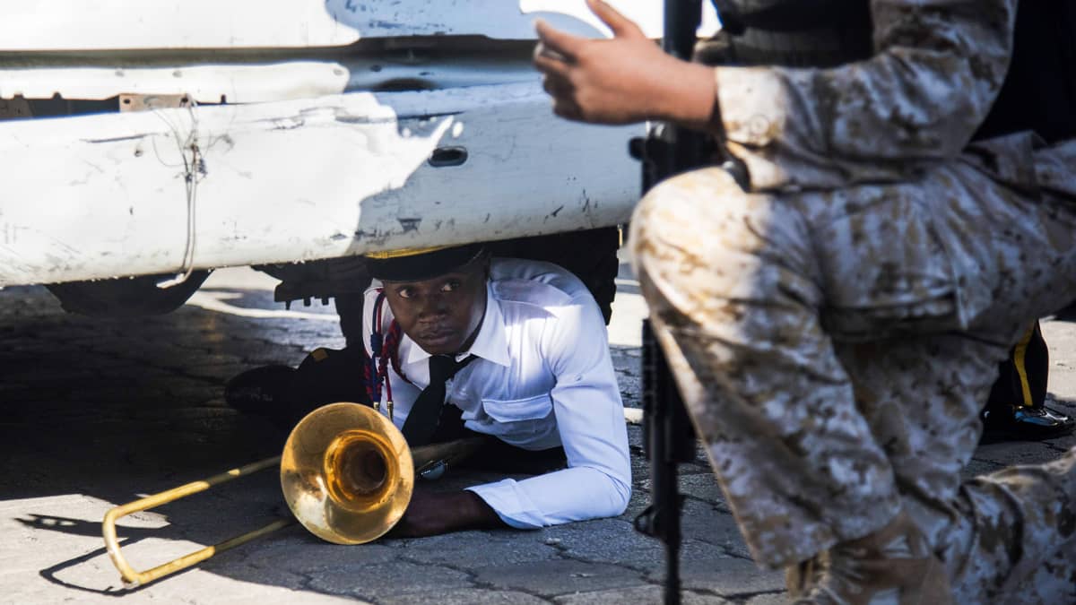 Haitin kansallispalatsin orkesterin jäsen suojautuneena auton alle soittimensa kanssa mellakan alkaessa.