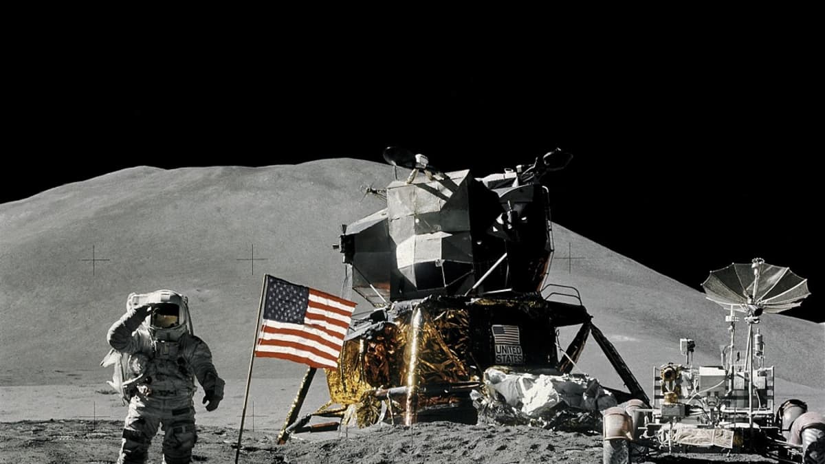 Astronautti Kuun pinnalla, edessä pajon jalanjälkiä, takana Yhdysvaltain lippu, laskeutuja ja mönkijä. Taustalla kukkula.