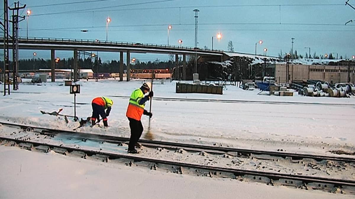 Kaksi miestä harjaa junanraiteita puhtaaksi lumesta.