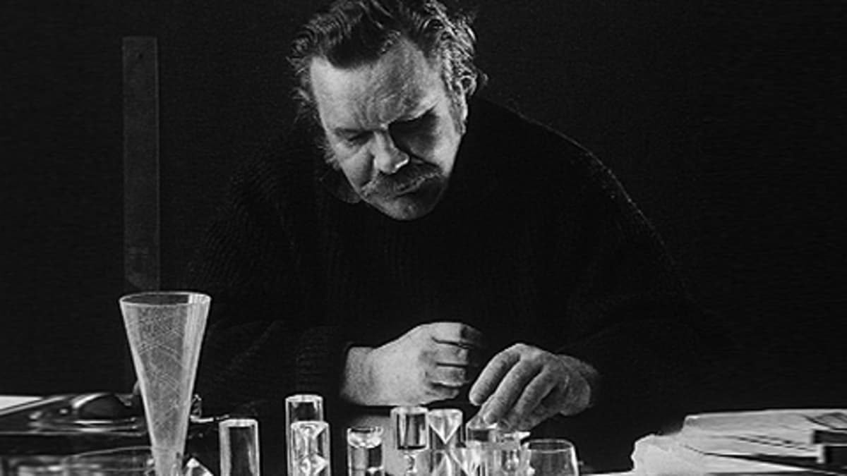 Arkistokuva Tapio Wirkkalasta, joka istuu työpöydän ääressä ja käsittelee lasiesineitä.