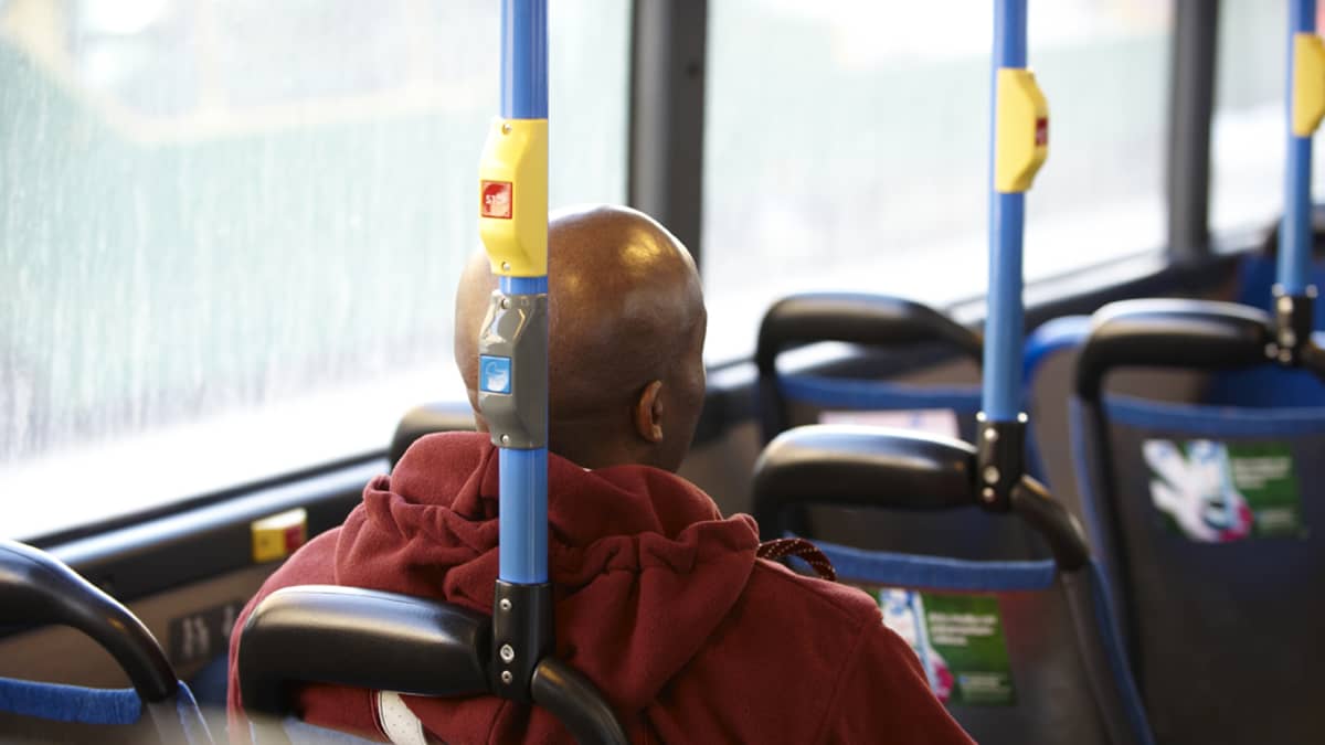 Tummaihoinen mies istuu bussin penkillä.