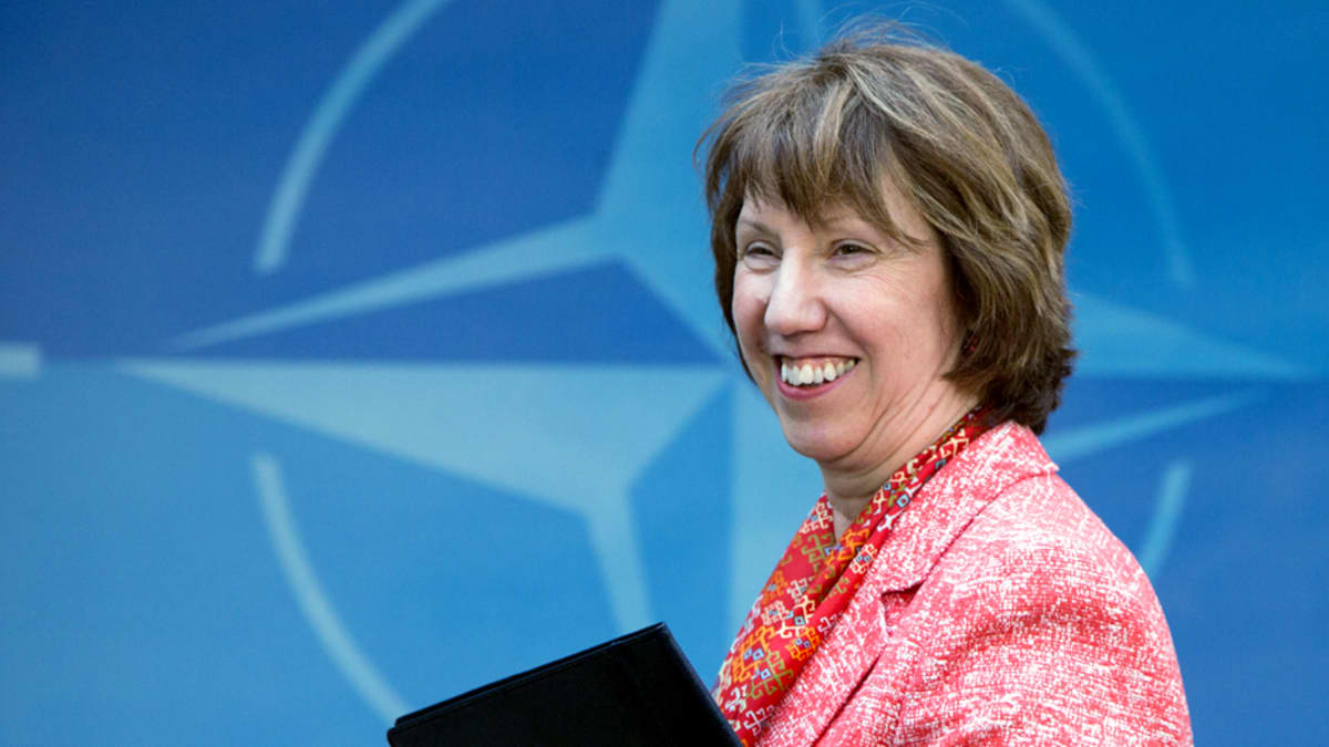 Catherine Ashton hymyilee, Naton logo taustalla.