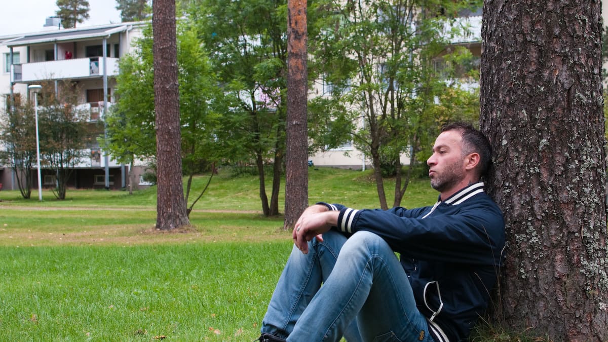 Turvapaikanhakija istuu nurmikolla ja nojaa puuhun.