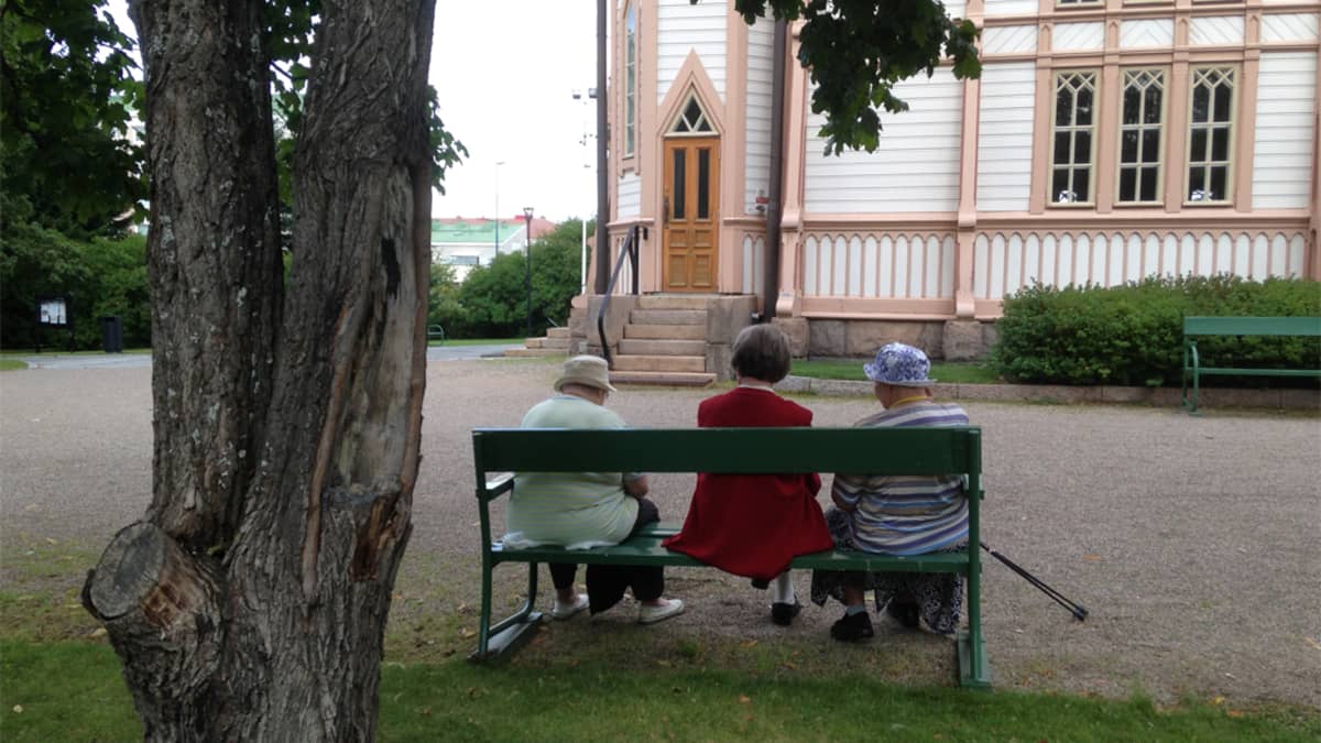 Kolme vanhaa naista istuu penkillä kirkon pihalla.