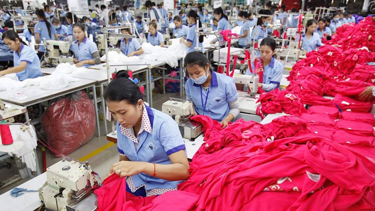 Naisia töissä tekstiilitehtaassa. Heidän ompelukoneidensa vierellä on suuria kasoja, joissa on t-paitoja ja kankaita.