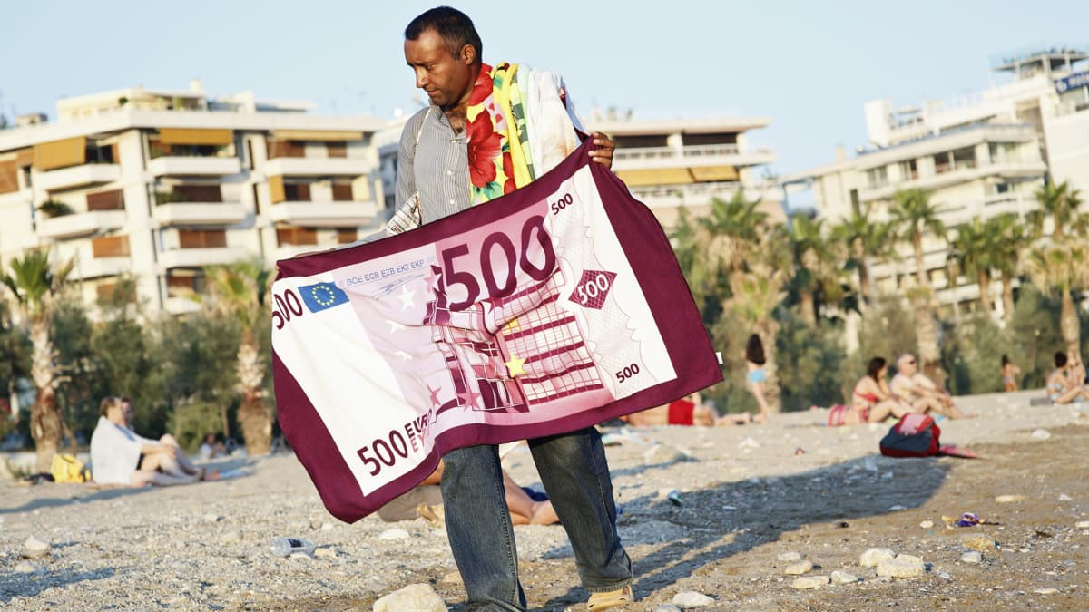 Mies kävelee Ateenan edustalla rannalla kädessään pyyhe, jossa on 500 euron setelin kuva.