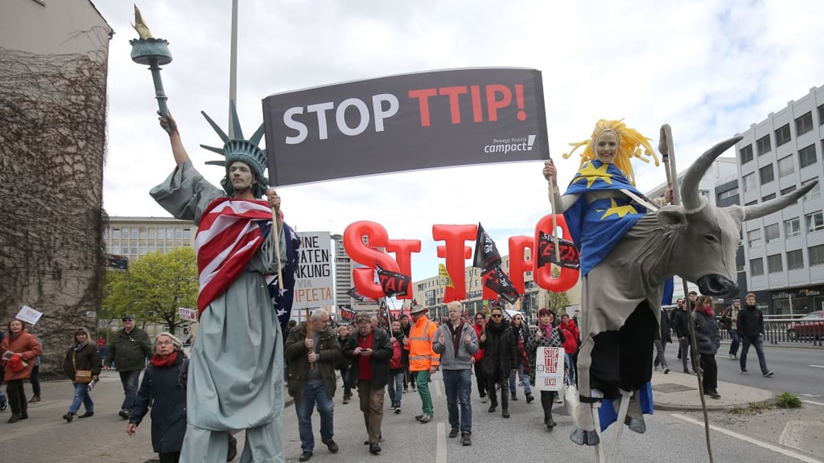 Vapaudenpatsaaksi pukeutunut mies ja EU:ta symboloiva nainen kantavat kylttiä, jossa lukee "Stopp TTIP!" Taustalla muita mielenosoittajia lippuineen. 
