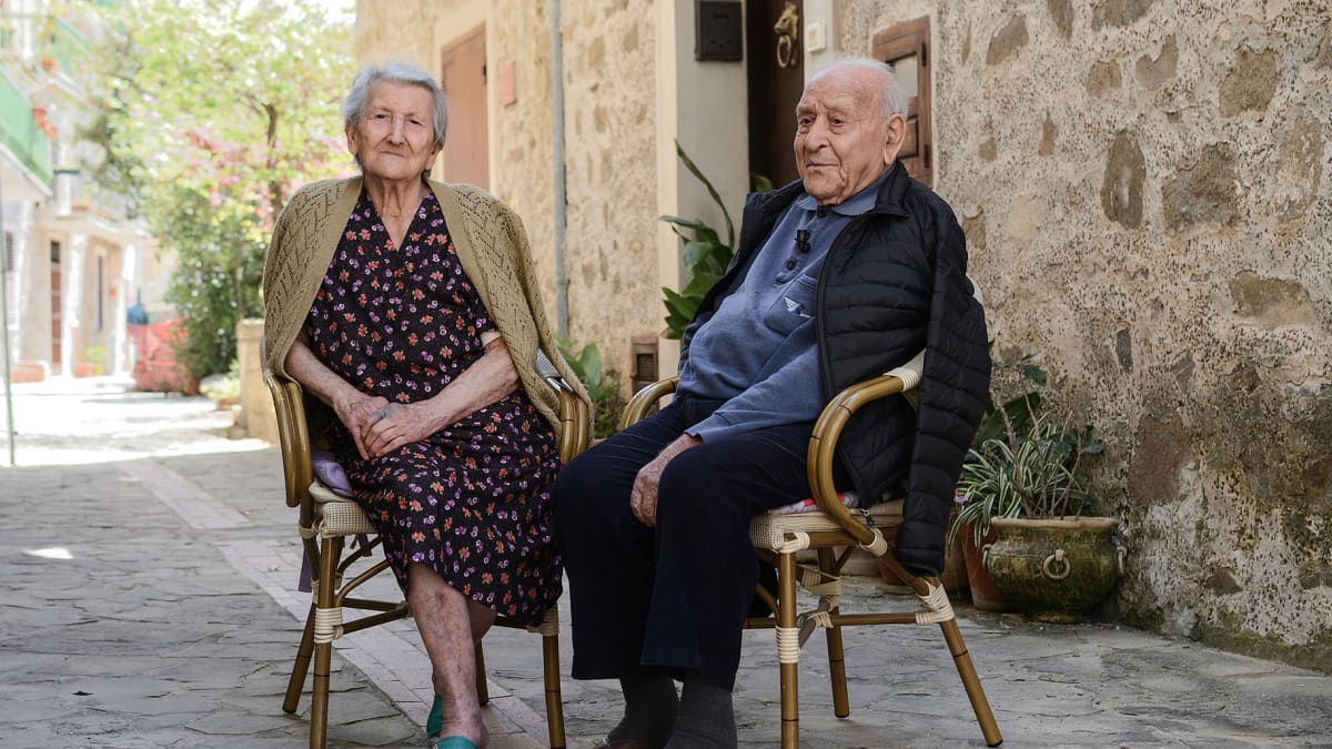 Amina Fedullo ja Antonio Vassallo nauttivat raikkaasta ulkoilmasta asuntonsa edustalla Acciarolin kylässä. Vanha pariskunta istuu tuoleilla.