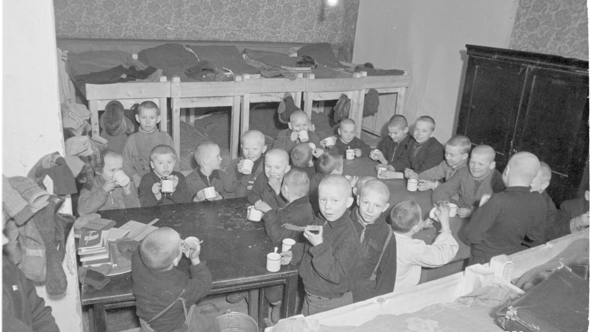 Noin kaksikymmentä poikalasta syömässä ahtaassa tilassa jossa on kerrossänkyjä.