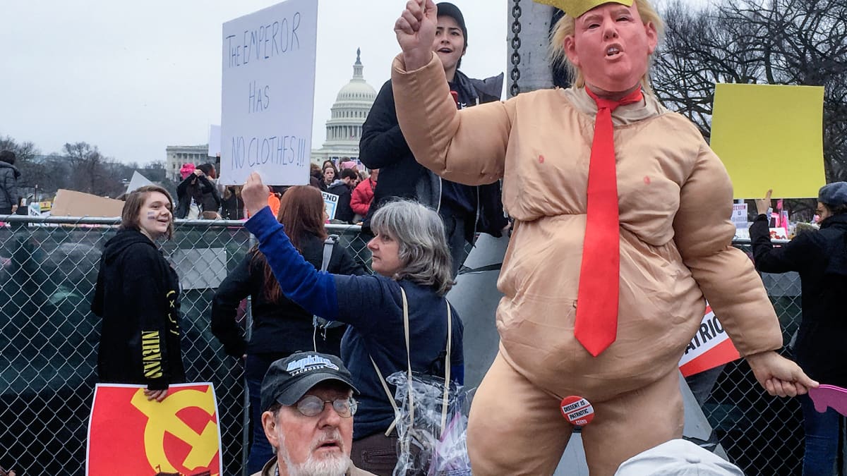 Henkilö on pukeutunut Donald Trump-naamariin ja pukuun, joka saa hänet näyttämään lihavalta ja alastomalta. Nainen pitää kylttiä, jossa lukee "The emperor has no clothes."