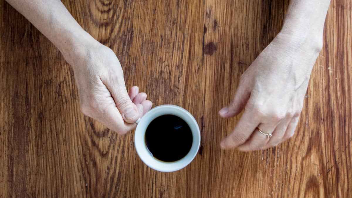 Henkilön kädet pöydällä, kädessä valkoinen kahvikuppi. 