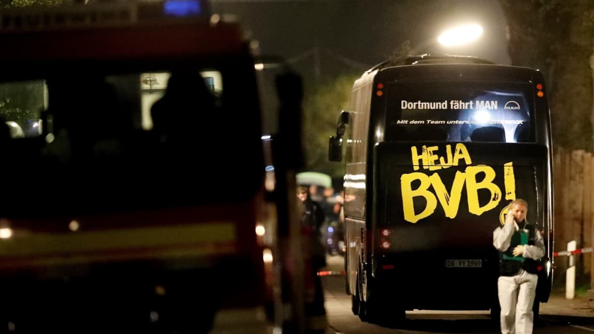 Kuva pommi-iskun kohteena olleesta jalkapallojoukkueen bussista. On ilta. Bussin takaosassa lukee suurin keltaisin kirjaimin "Heja BVB!" Etualalla kulkee nainen ja puhuu puhelimeen.