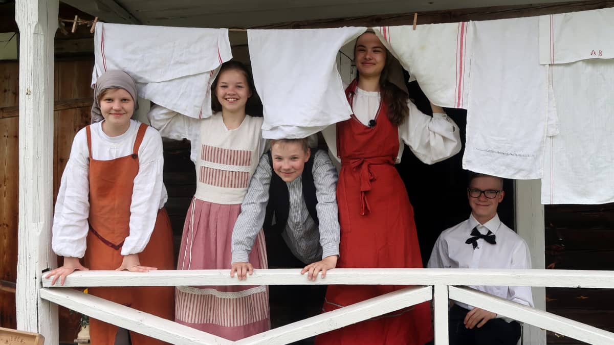 Kuvassa nuoria 1800-luvun vaatteissa pyykinarun takana.