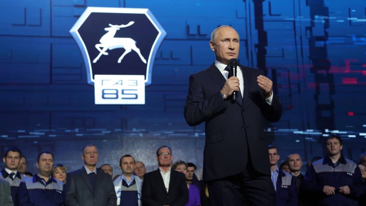 Putin puhuu lavalla mikrofoniin. Taustalla näkyy joukko tehtaan työntekijöitä ja GAZ-yhtiön logo.