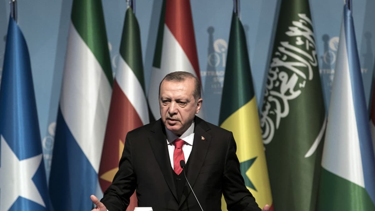 Erdoğan seisoo puhujankorokkeen takana ja puhuu mikrofoniin. Hänellä on tumma puku, liivit ja valkoinen kauluspaita sekä punainen kravatti. Taustalla on kokouksen Islamilaisen yhteistyöjärjestön jäsenmaiden lippuja.