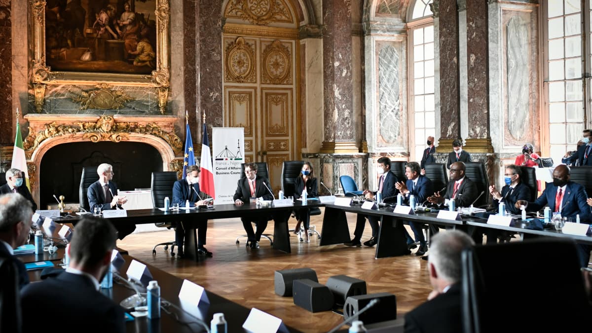 Iso neliönmuotoinen pöytä Versailles'n juhlasalissa, liikemiehiä pöydän ympärillä, Macron keskellä.