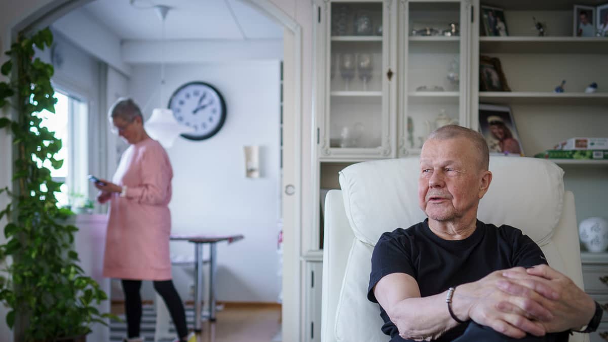 Harri Lyytikäinen istuu nojatuolissa olohuoneessaan. Taustalla elämänkumppani Maija Abrahamsson.