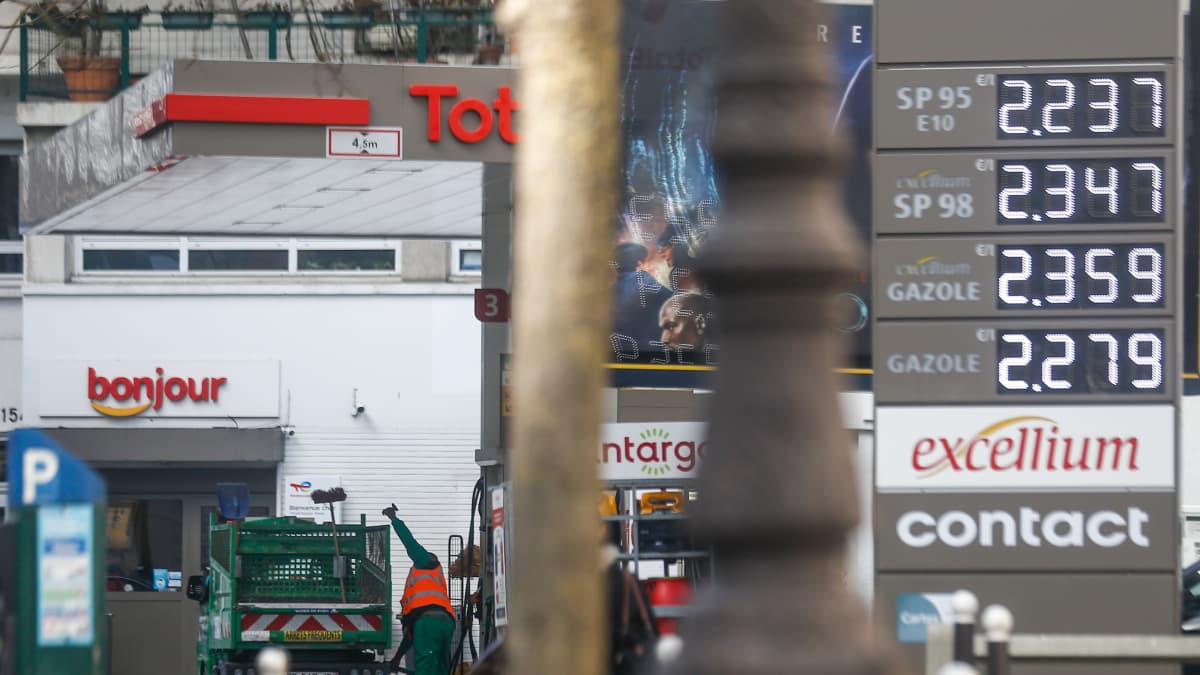 Yleiskuva huoltoasemalta harmaana päivänä, taustalla mies huomioliiveissä tankkaa. Hintataulu näyttää esimerkiksi 95-oktaanisen bensan hinnaksi 2,237 euroa.