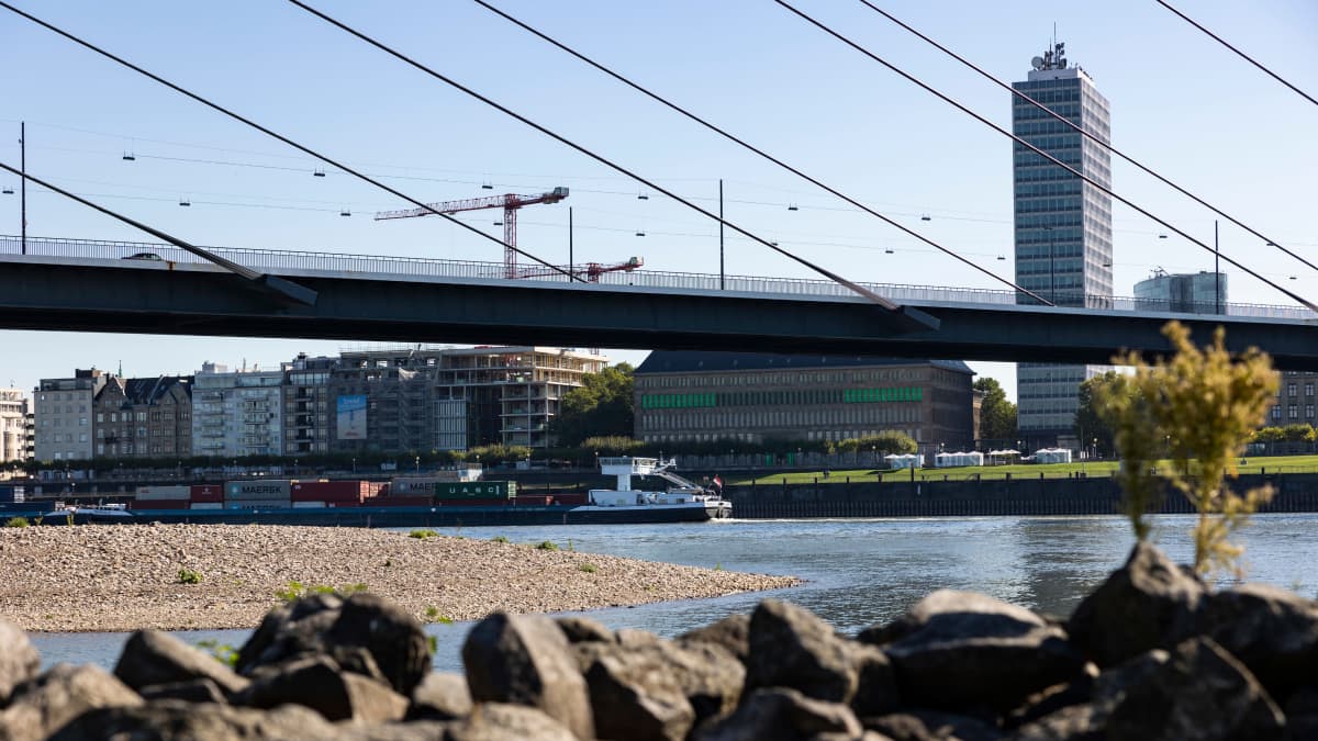 Etualalla kivenjärkäleitä ja hiekkaa, taustalla erottuu joen vettä, silta ja Düsseldorfin silhouetti.