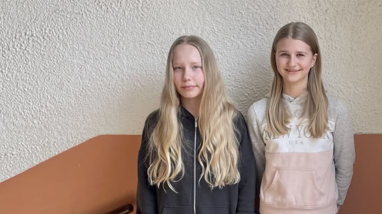 Kaksi kuudesluokkalaista tyttöä seisoo vierekkäin koulun käytävällä ja katsoo kameraan. 