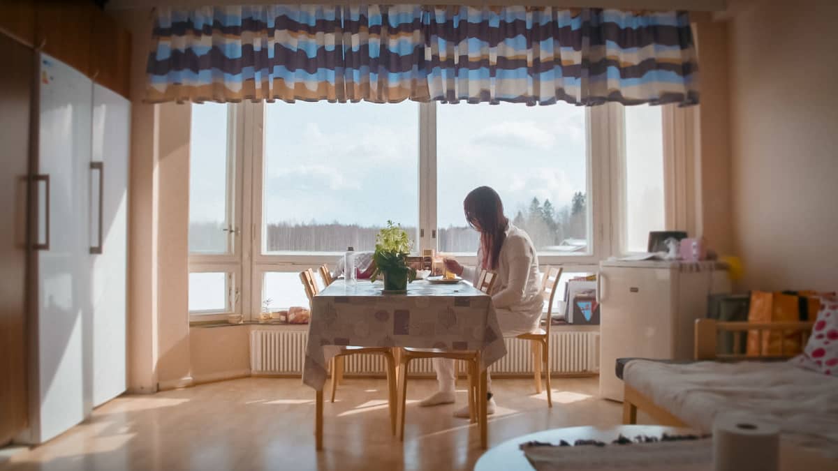 Nainen istuu ruokapöydän ääressä ikkunan vieressä syömässä.