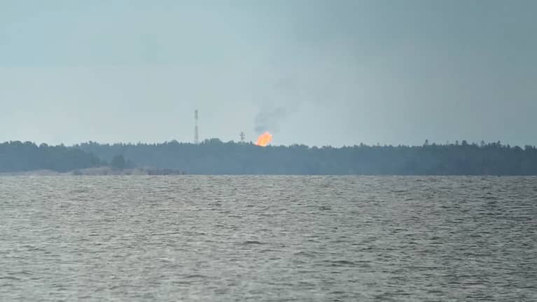 Meri, jonka takana on rannikolla metsä. Metsän takaa nousee tulenlieska.