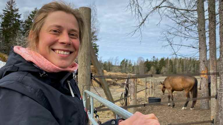 En kvinna som står vid hästhagen och tittar in i kameran. 