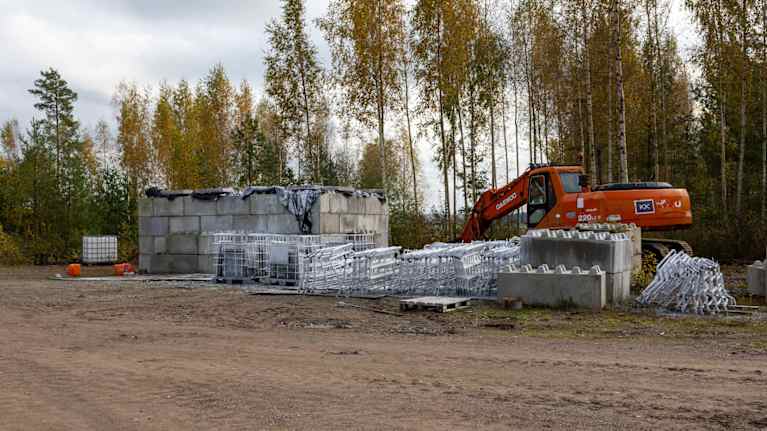 En stor behållare av betong med en orange grävmaskin bakom och vita metallstängselbitar framför.