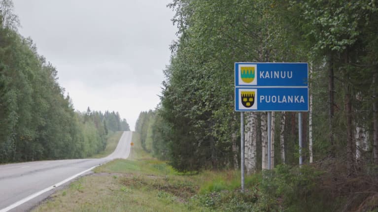 Tyhjä maantie kesällä, tien vieressä kyltti, jossa lukee 'Kainuu' ja 'Puolanka'.