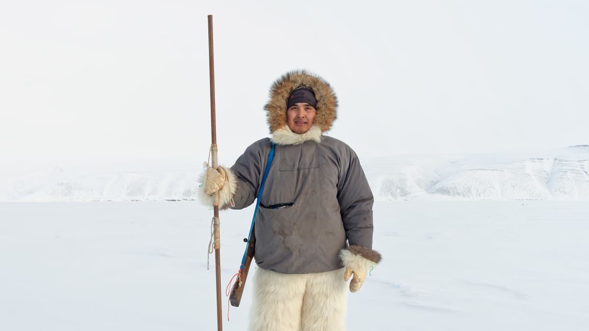 Jääkarhuhousuihin pukeutunut mies seisoo harppuuna kädessä jääkentällä.