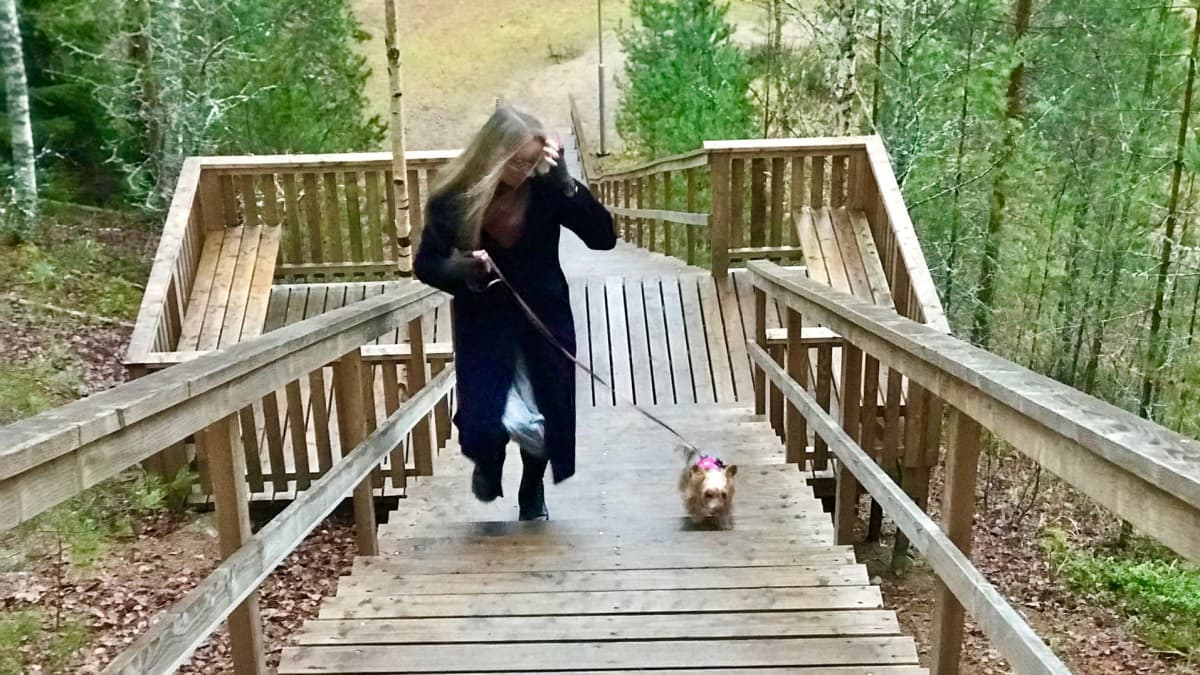 Vaalea, pitkätukkainen nainen lilassa pitkässä takissa juoksee ylös kuntoportaita pienen koiran kanssa