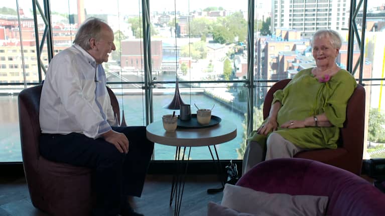 Esko and Eila Roine are sitting on either side of a small round table chatting. Esko is wearing a white shirt, and Eila is wearing a greenish tunic and gray pants. 